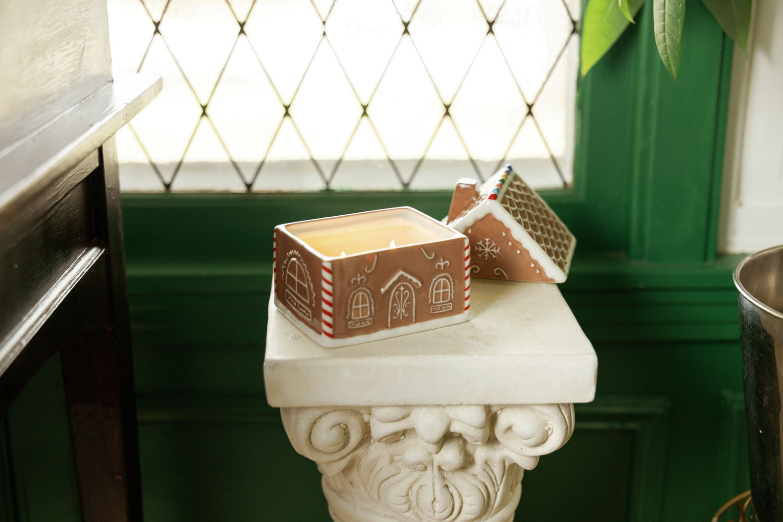 A gingerbread-themed candle in a ceramic container with a house design, displayed on a white stone pedestal in front of a green paneled wall and diamond-patterned window