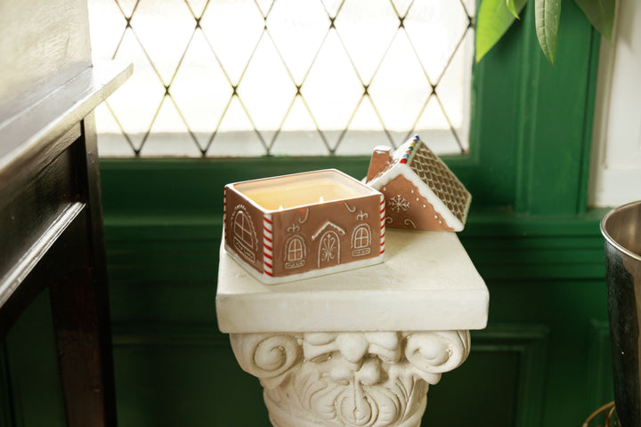 A gingerbread-themed candle in a ceramic container with a house design, displayed on a white stone pedestal in front of a green paneled wall and diamond-patterned window
