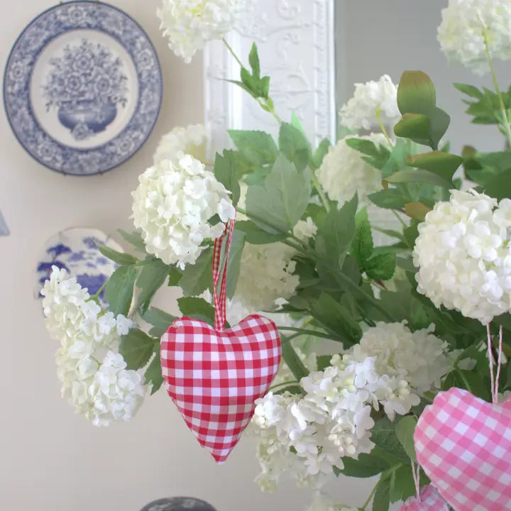 A red and white gingham heart-shaped fabric ornament hanging from a bouquet of white hydrangea flowers, with a matching pink gingham heart ornament partially visible in the foreground.