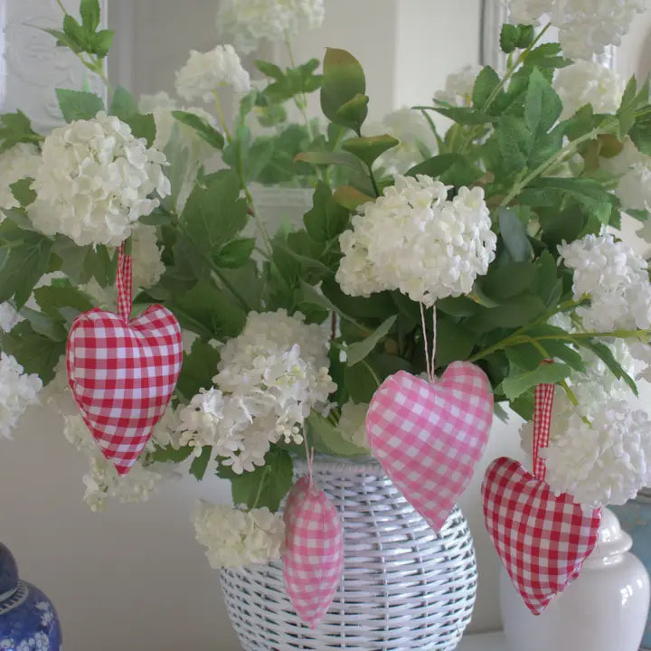 Four pink and red gingham fabric heart ornaments hang from a white wicker vase filled with white flowers and green leaves.