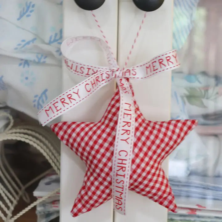 A red and white gingham star-shaped fabric ornament hanging from a white door, tied with a white ribbon printed with 'MERRY CHRISTMAS' in red.