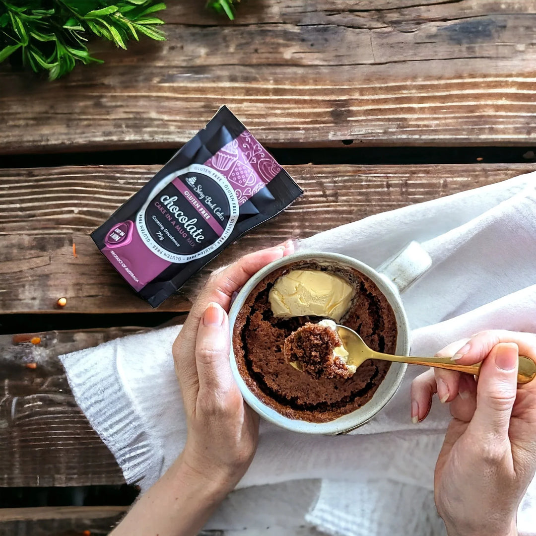 Chocolate mug cake with a scoop taken out, held by hands over a wooden surface with a package of chocolate mix.