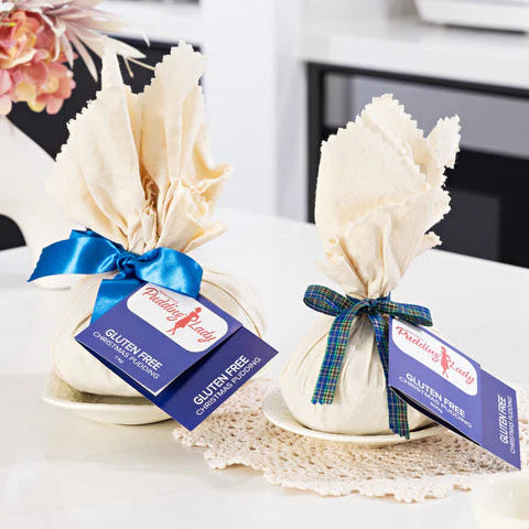 Two Christmas puddings wrapped in calico and tied with blue ribbons, each with a blue hangtag reading Gluten Free. Styled on a white table with a vase in the background.