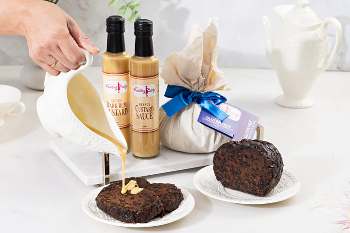 A hand pours creamy custard sauce from a white pitcher onto chocolate scones on a white plate, with two bottles of custard sauce, a wrapped gift tied with a blue ribbon, and another scone on a white table.