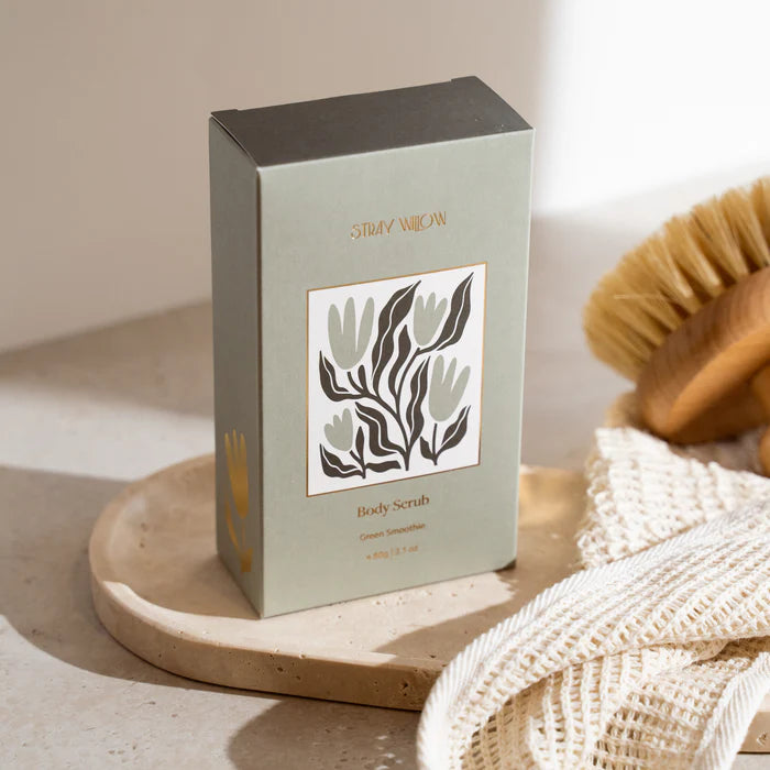 A green box of Stray Willow body scrub on a marble surface with a body scrub &amp; loofah.