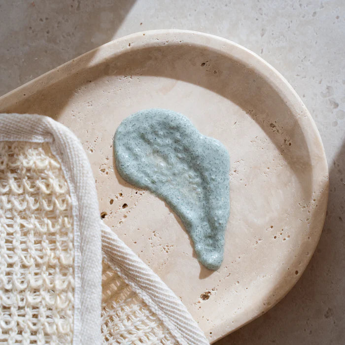 Beige ceramic plate with a dollop of green body scrub and a loofah on a neutral background