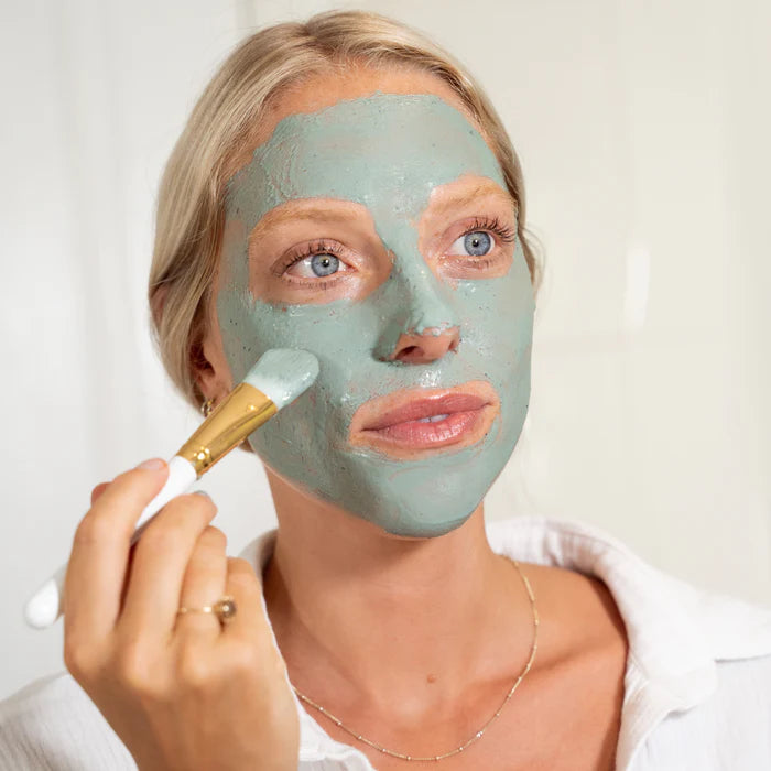 Woman applying a green facial mask with a brush on a white background