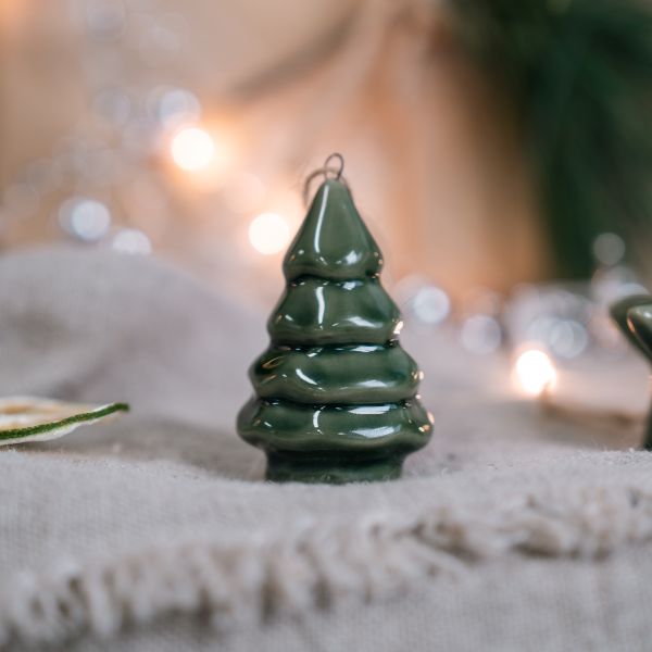A small glossy dark green Christmas tree ornament with a wire hanger loop at the top, standing on a textured beige surface with warm blurred lights in the background.