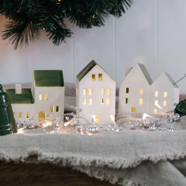A set of small white ceramic house-shaped ornaments with green roofs and warm yellow window lights, arranged on a textured beige blanket with silver beads and fairy lights, with a Christmas tree branch in the background.