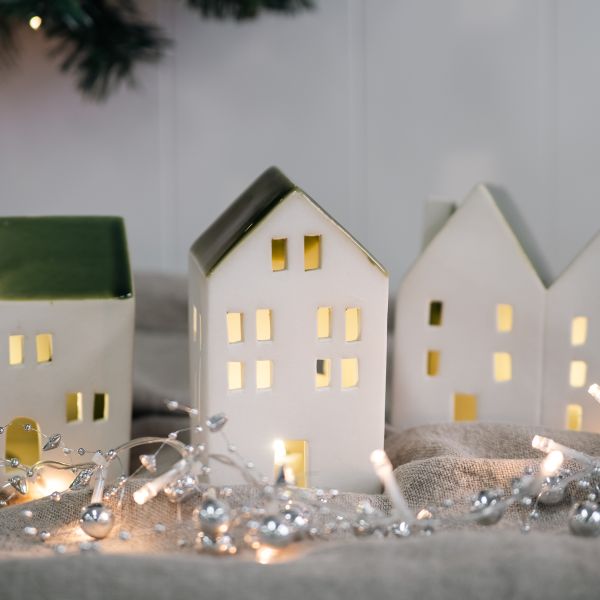 A set of small white ceramic house-shaped candle holders with warm yellow glowing lights inside, styled on a textured fabric surface with silver tinsel and fairy lights in a cozy indoor setting.