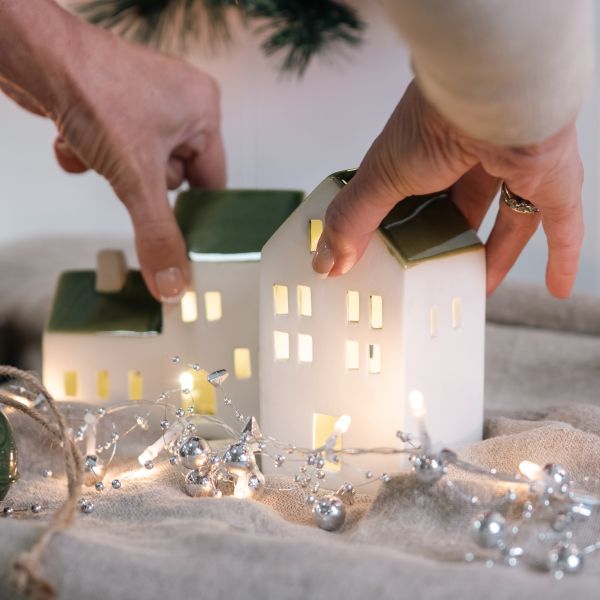 Two hands are holding a small white ceramic Christmas village house ornament with a green roof and glowing warm yellow windows, alongside another similar house and silver holiday lights on a beige fabric surface.