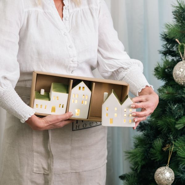 A person in a white blouse and beige apron holds a box of ceramic house-shaped ornaments next to a decorated Christmas tree. The houses are cream-colored with green roofs and glowing yellow window lights.