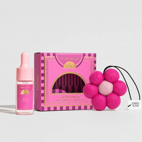 Three Suns air freshener set featuring a pink dropper bottle, a pink and gold patterned box, and a pink felt flower-shaped diffuser with black cords on a light gray background.