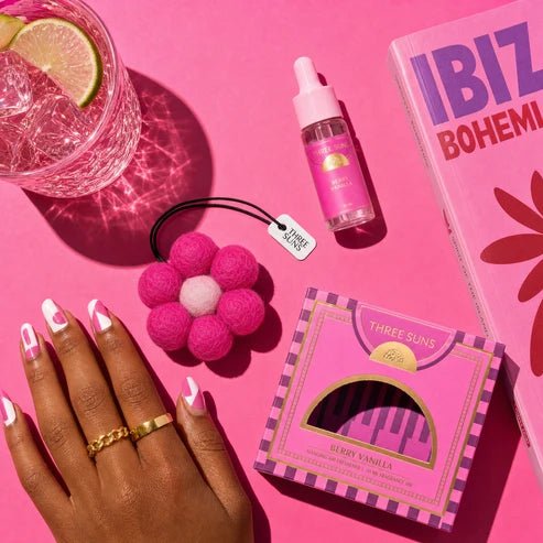 A styled flat-lay featuring a pink and purple striped box of Three Suns Berry Vanilla nail wraps, a pink flower-shaped hair tie, a pink dropper bottle, and a pink book, arranged around a hand with white and pink manicured nails on a bright pink background.