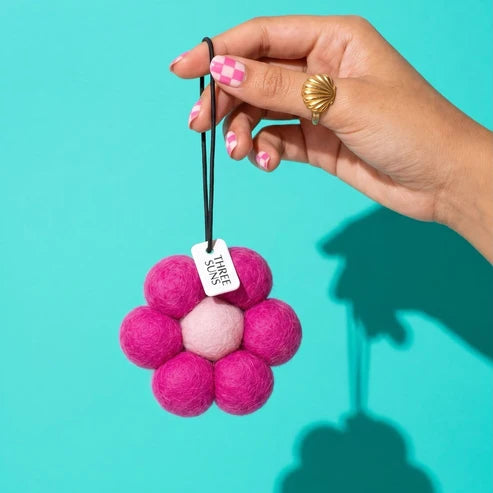 A hand with pink checkered nail polish and a gold ring holds a pink felt flower-shaped hanging ornament with a small white tag against a bright turquoise background.
