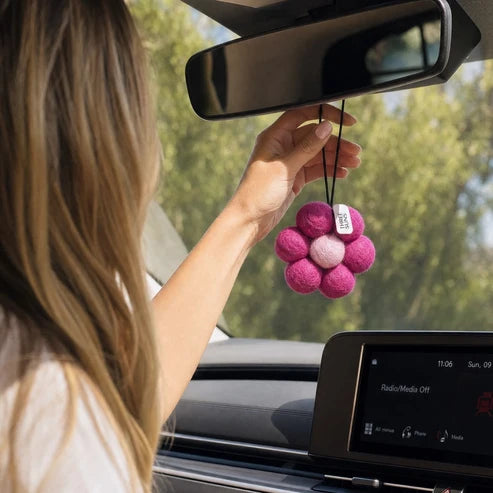 A person hanging a pink flower-shaped car air freshener with a small white tag from the rearview mirror inside a car.