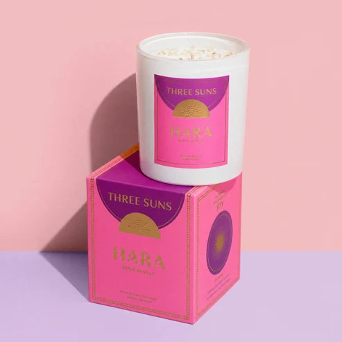 A white glass candle jar with a pink and purple label sits on top of its matching pink and purple box. The product is branded Three Suns and named Hara, with &