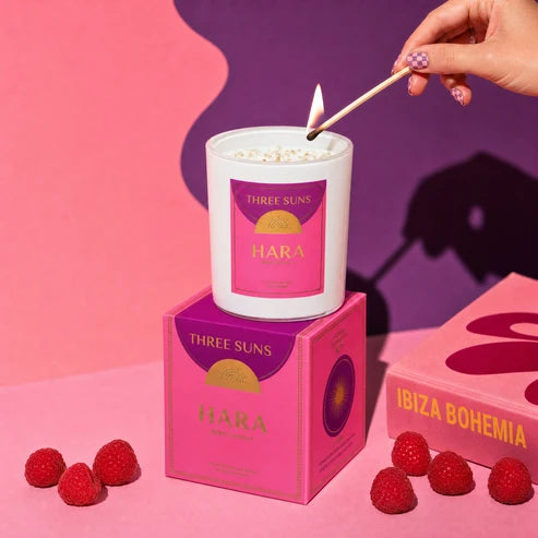 A white candle labeled HARA by Three Suns is lit with a wooden match, resting on top of its pink and purple product box. The scene is styled with fresh raspberries and a pink book, set against a vibrant pink and purple background.
