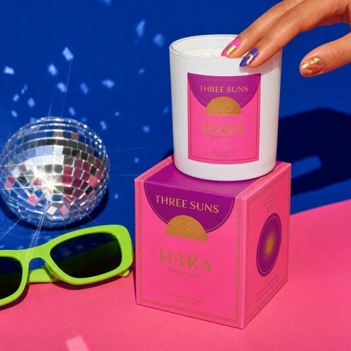 A white candle jar with a pink and purple label sits on top of its matching pink box. A hand with metallic nail polish rests on the candle, and bright green sunglasses and a disco ball appear at the sides against a vivid blue background with confetti-like streaks.