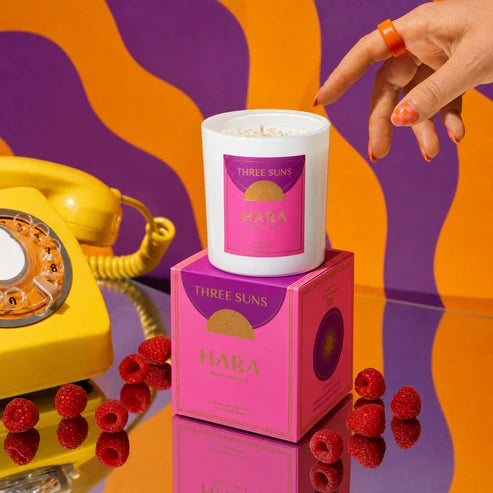 A Three Suns Hara Spice Melia scented candle in a white jar sits on top of its pink and purple packaging box, styled with fresh raspberries on a reflective surface. A hand with orange nail polish points toward the candle, and a retro yellow rotary phone is visible in the background against a wavy orange and purple backdrop.