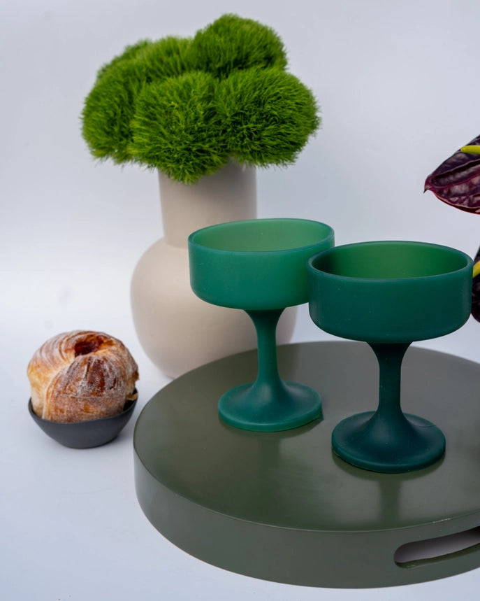 Two green silicone cocktail glasses displayed on a round green tray, with a beige vase of fluffy green foliage and a pastry in the background on a white studio surface.