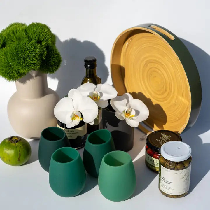 A styled product arrangement on a white surface featuring matte green stemless wine glasses, a large round wooden serving tray with green edging, a beige ceramic vase with green moss, white orchids in a small black vase, a dark bottle, a green fruit, and two jars.