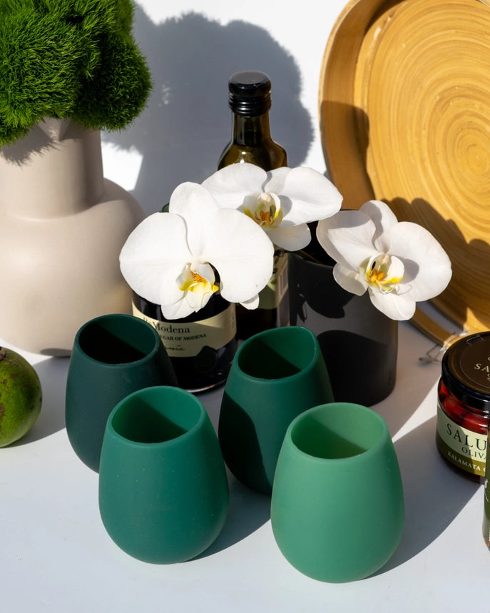 Four matte stemless silicone wine glasses in shades of green arranged on a white surface, styled with white orchids, a dark bottle, a beige vase with greenery, and a round wooden plate in bright sunlight.