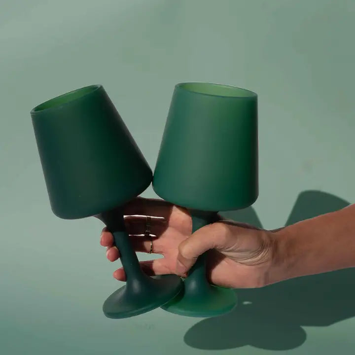Two dark green silicone unbreakable glass goblets held together by a hand against a solid sage green background.