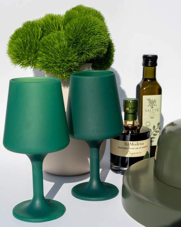 Two matte green silicone wine glasses, styled on a white surface with a potted green plant, olive oil bottles, and a green ceramic object in the background.