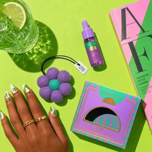 A styled flat-lay featuring a purple flower-shaped felt freshener with a green center and a black elastic band, shown next to its purple and green patterned packaging box. The scene is set on a bright lime green background alongside a hand with green and white manicured nails, a glass of water with a lime slice, and a pink book.