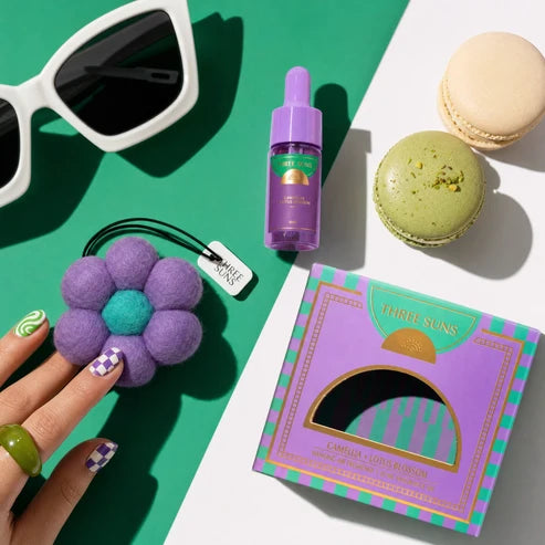 Purple product with packaging, sunglasses, macaron, and hand holding a purple flower-shaped item on a green and white background.