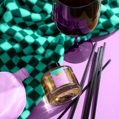 A glass reed diffuser bottle with a pink label and black cap sits on a lavender surface beside several black reeds. A purple wine glass, a pink balloon, and a green-and-black checkered blanket are arranged around it in a styled flat-lay.