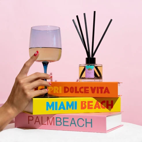 Hand holding a glass of wine with books and a diffuser on a pink background