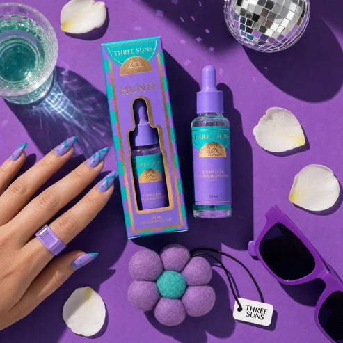 Three Suns Hunti fragrance oil bottle and its packaging box, styled in a flat lay on a purple background with a hand, sunglasses, a disco ball, and flower accessories.