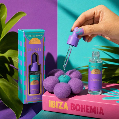 A hand with purple nail polish holds a purple dropper above a small bottle and its matching box. The product packaging and bottle label read Three Suns Hunti, with a pink block labeled Ibiza Bohemia in the foreground and purple and teal background panels with green leaves.