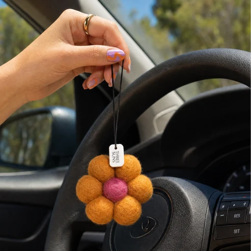 A hand holds a small felt flower car air freshener with orange petals and a pink center, hanging near the steering wheel inside a car.