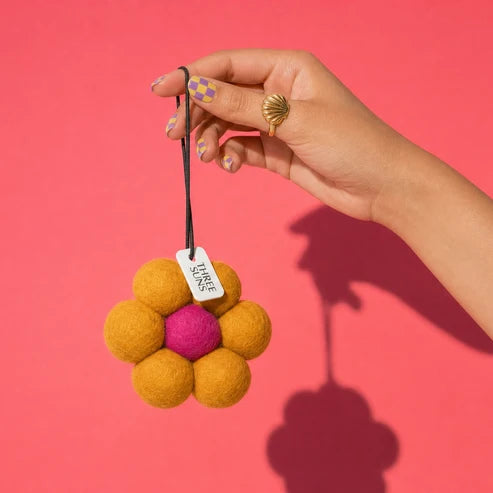 A hand holds a small felt flower ornament against a solid pink background. The ornament has a bright pink center and six yellow felt petals, attached to a black string with a white tag that reads &