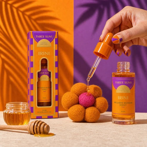Three Suns Irini fragrance oil, featuring an open dropper bottle, its orange and purple packaging box, a small glass jar of honey, and a honey dipper on a textured beige surface. A hand with purple and orange nail polish is shown holding the dropper cap over the bottle.