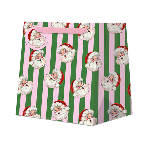 A festive Christmas gift bag featuring a repeating pattern of cartoon Santa Claus faces over vertical stripes of green and pink, with a pink handle and a matching pink hanging tag.