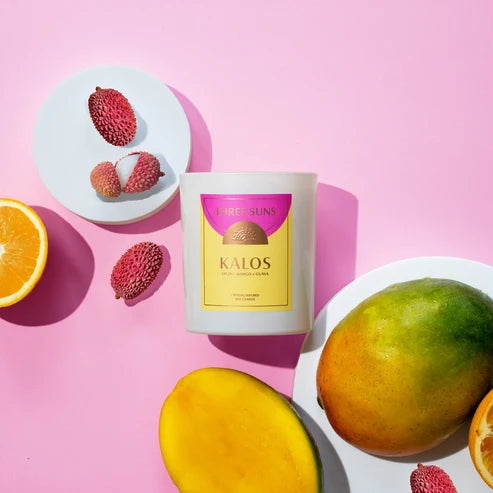A white candle jar labeled THREE SUNS KALOS sits on a pink background with fruit props including lychee fruits, a mango, and orange slices on white plates.