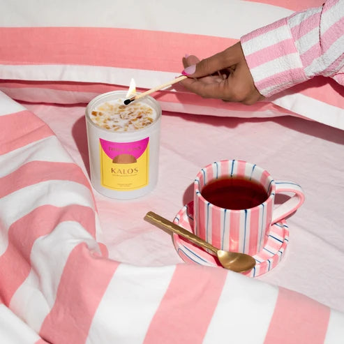 Candle being lit next to a cup of tea on a pink and white striped blanket, with &