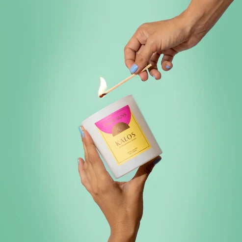 A hand holds a white candle jar with a pink and yellow label while another hand ignites it with a lit wooden match against a solid mint green background. The brand name &
