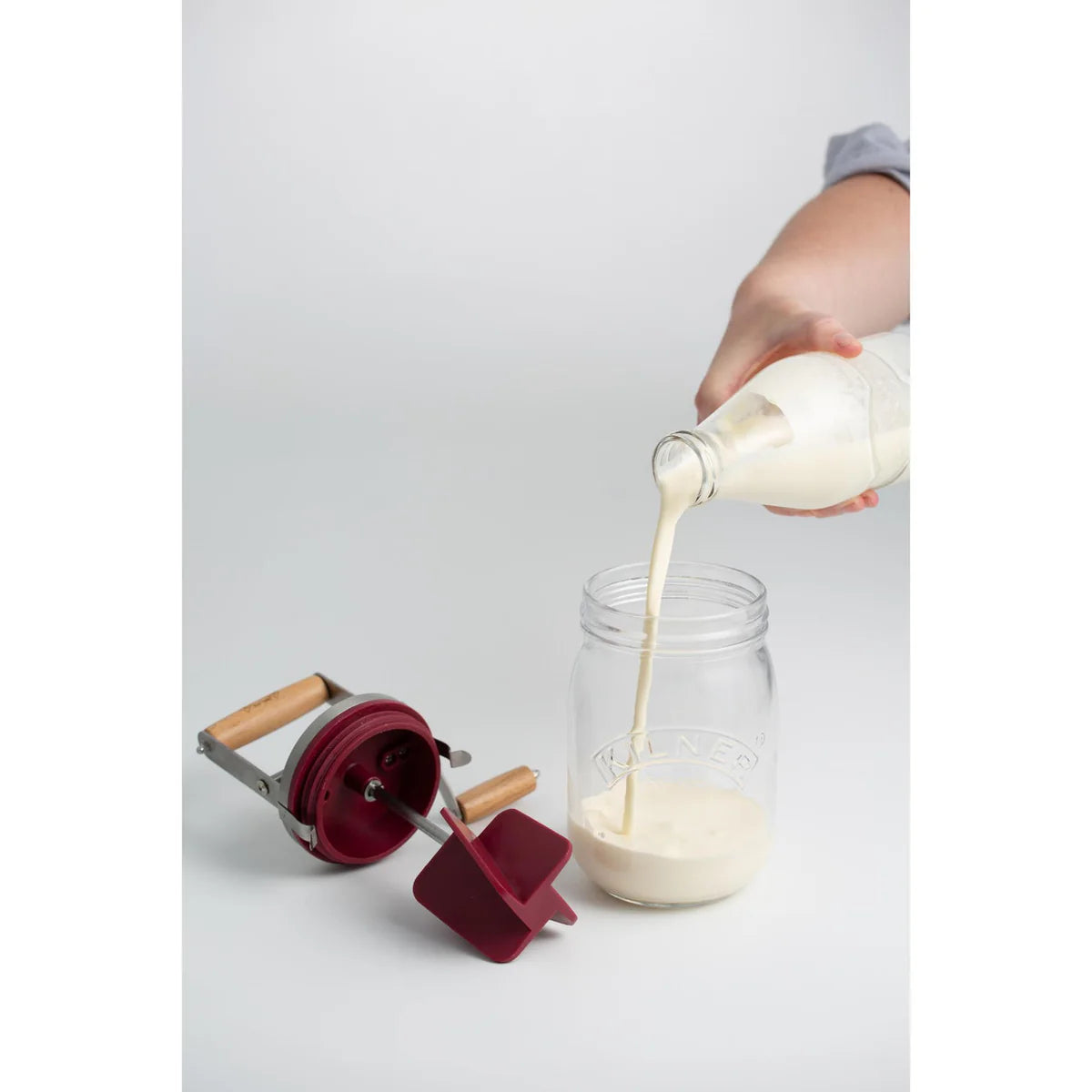 Person pouring liquid from a bottle into a jar with a red and silver tool on a light gray background