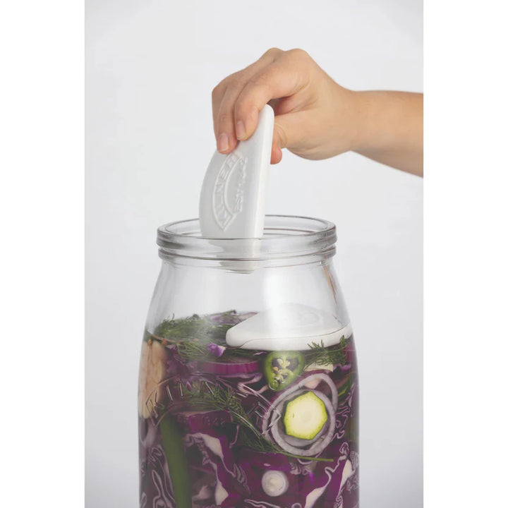 Hand placing a white disc on a jar with vegetables inside, against a white background