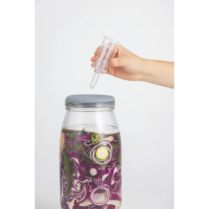 Glass fermentation jar with a hand holding a clear dropper over it on a white background