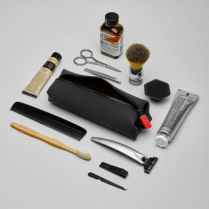Black zippered toiletry pouch on a white background, surrounded by toiletry items including razor, nail file, toothbrush, shaving brush, beard oil