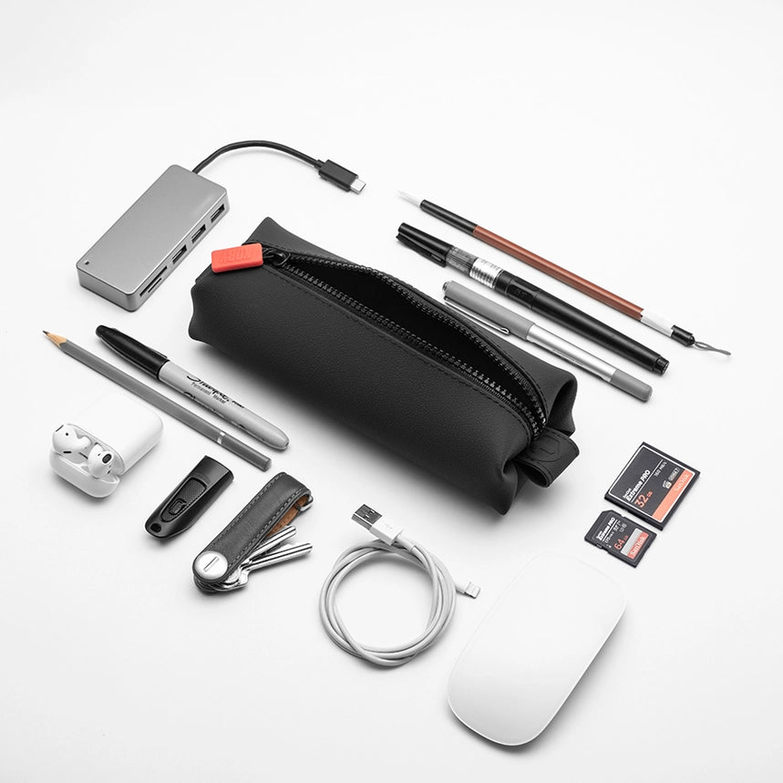 Black zippered toiletry pouch on a white background, surrounded by tech items including mouse, cable, data port, ear buds