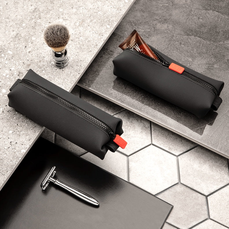 Two black zippered toiletry pouches on grey tiles with a shaving brush and razor