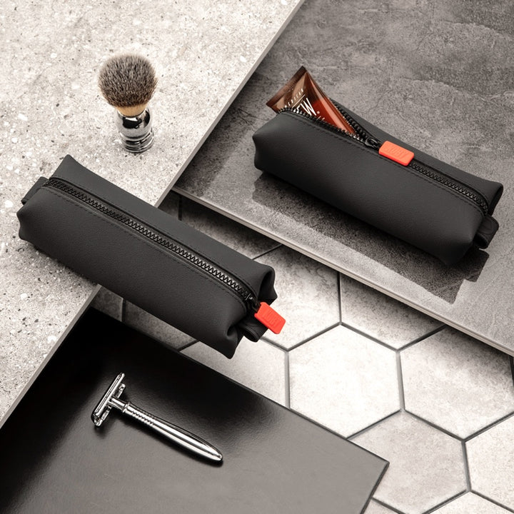 Two black zippered toiletry pouches on grey tiles with a shaving brush and razor