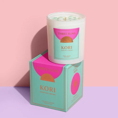 A white glass candle jar labeled 'THREE SUNS KORI' sits on top of its matching mint-green and pink box against a pink and lavender studio background.